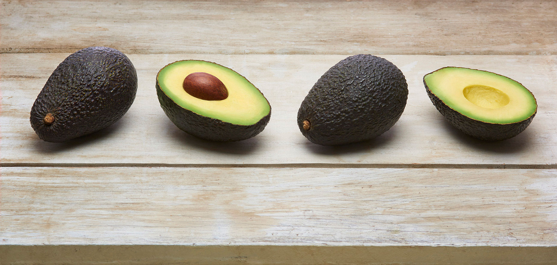 Two-whole-and-one-cut-hass-avocad-on-rustic-white-wood-table-top-by-Joe-Atlas-food-photographer.